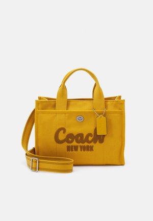 Bolso de lona amarillo con asas cortas, correa de hombro ajustable, cierre delantero de giro y logo "Coach New York" en el frente.