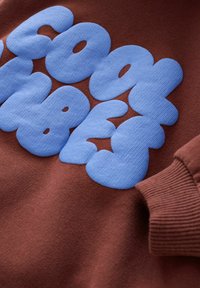 Sweatshirt marron avec de grandes lettres en bulle bleues, douces, inscrites "COOL VIBES". Tissu texturé avec des poignets côtelés.