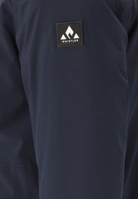 Mörkblå jacka med en slät textur, utrustad med en svart rektangulär logopatch med vit bergdesign på bröstet.