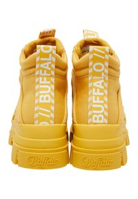 Zapatillas altas de tela amarilla con suelas de goma gruesas, que presentan una marca blanca y franjas en el talón y en las lengüetas.