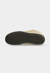 Semelle d'une basket beige montrant une bande de roulement noire avec un logo en forme de chat au centre.