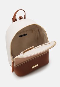 Petit sac à dos avec partie supérieure crème, base et sangle marron, fermetures éclair dorées, et compartiment principal ouvert montrant une poche intérieure zippée portant l'étiquette Anna Field.