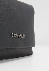 Coin de sac à main en cuir noir texturé avec logo Calvin Klein argenté sur une surface blanche et un fond gris clair uni.