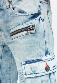 Light blue denim pants with a washed look, featuring a zipped pocket, ribbed accents on the thighs, and a logo patch.