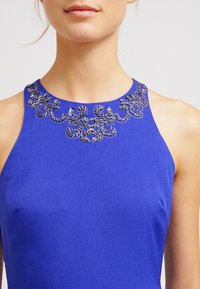 Královsky modré šaty bez rukávů s vysokým výstřihem zdobeným složitými stříbrnými ozdobami podél límce. Hladká textura látky.