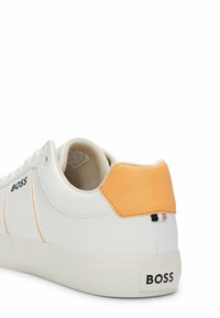 Zapatilla blanca con acento naranja en el talón, suela de goma y costuras en contraste. Presenta un logo en el lateral y forro de tejido texturizado.
