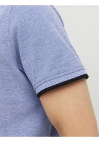 Kortärmad ljusblå strukturerad skjorta med svart kant på ärmarnas fåll, där en del av en persons arm syns mot en enfärgad bakgrund.