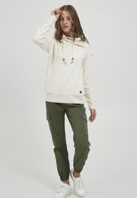 Crème hoodie met trekkoord en houten accenten, gecombineerd met olijfgroene cargobroek. Model staat voor een neutrale achtergrond.