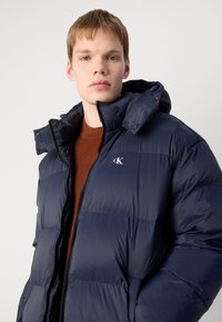 Marineblå puffjakke med glidelås, høy krage og merkevarelogo. Laget av glatt, quiltet stoff med en blank finish.