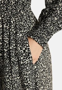 Hand in seitlicher Tasche eines schwarz-weißen Leopardenmuster-Kleids mit gerafftem Bund und gesmoktem Ärmelabschluss.