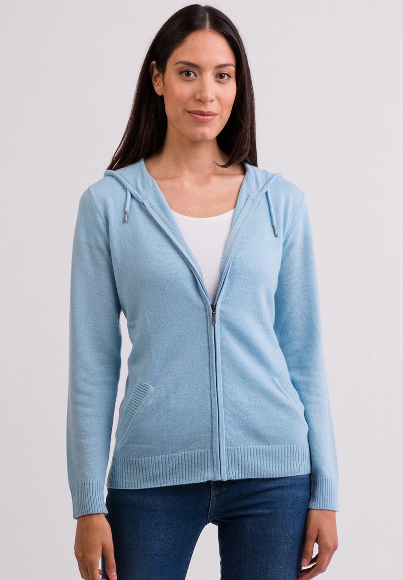 Ljusblå zip-hoodie gjord av mjukt material med huva, framfickor och ribbade ärmar. Har en slät textur och minimalistisk design.