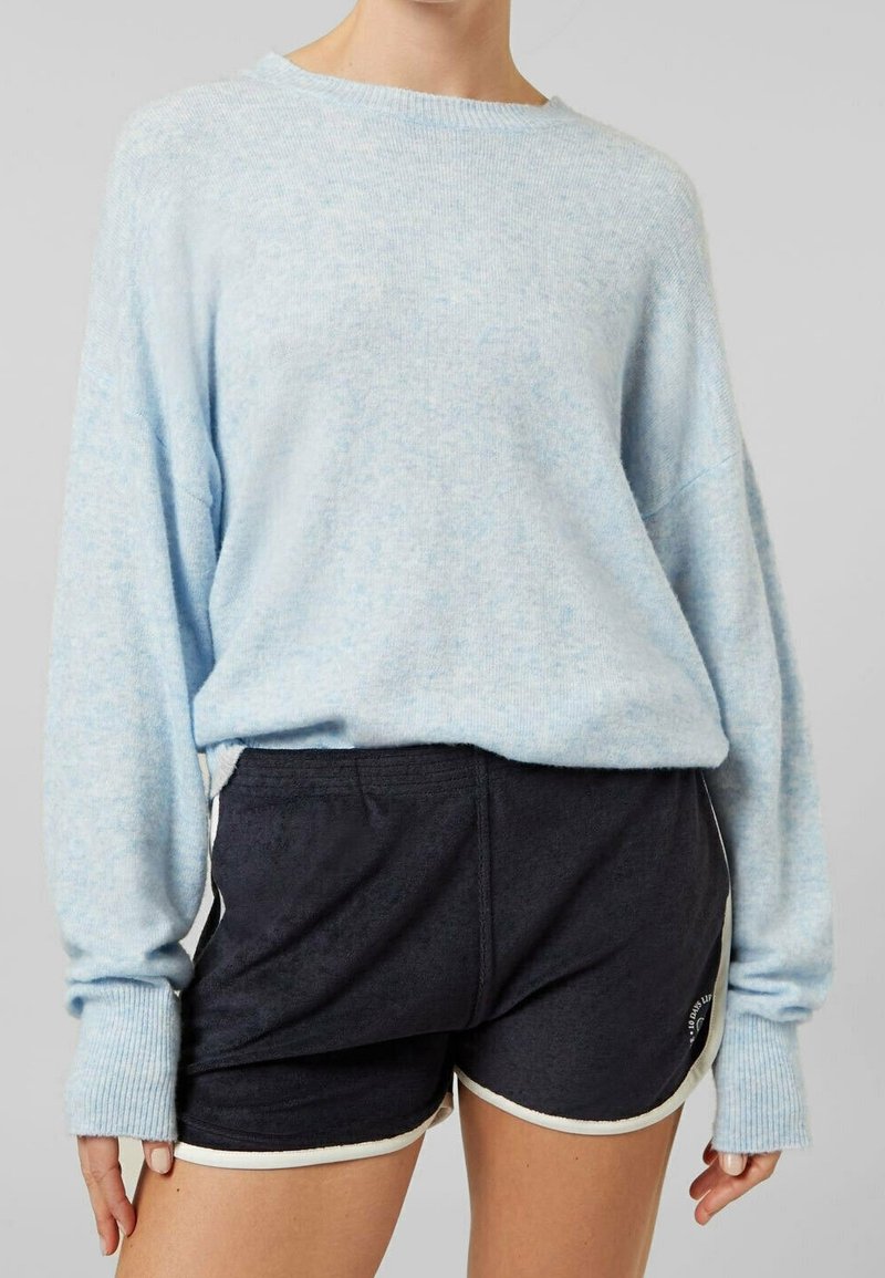 Personne portant un pull à manches longues bleu clair et un short de sport noir avec une bordure blanche sur un fond gris uni.