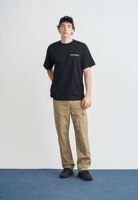 Camiseta negra de algodón con logo blanco, combinada con pantalones cargo beige con múltiples bolsillos. Complementado con zapatillas negras y una gorra.