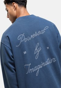 Felpa blu navy con maniche lunghe, caratterizzata da un testo ricamato bianco sulla schiena che recita "Powered by Imagination" e "20 11."