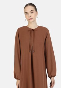 Bruine jurk met lange mouwen en ronde halslijn, met een strikdetail aan de voorkant en elastische manchetten, gemaakt van lichte, textuurstof.