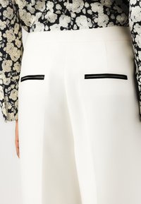 Pantaloni bianchi su misura con piping neri sulle tasche posteriori. Il tessuto ha una sottile trama, che si abbina a una maglia stampata floreale in bianco e nero.