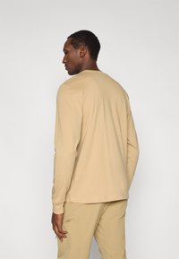 Μπεζ πουκάμισο με μακριά μανίκια και στρογγυλή λαιμόκοψη, κατασκευασμένο από απαλό ύφασμα, φαίνεται από πίσω συνοδευόμενο από μπεζ παντελόνια το ίδιο χρώμα. Καθαρός σχεδιασμός, χωρίς σχέδια.