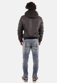 Giacca di pelle marrone con cappuccio, caratterizzata da dettagli cuciti, abbinata a jeans blu chiaro con effetto sbiadito e motivo splatter.