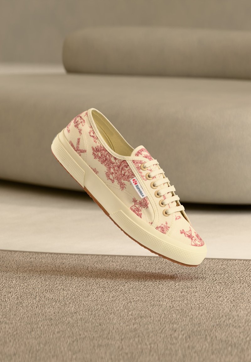 Beige snøresneakers med rødt toile-mønster og hvit såle, merket "Superga," som svever over en beige teppe med tekstur i et nøytralt rom.