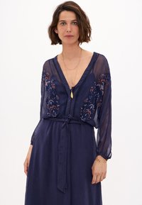 Robe bleu marine avec de longues manches transparentes, détails floraux brodés sur le corsage, décolleté en V profond et ceinture en tissu à la taille.