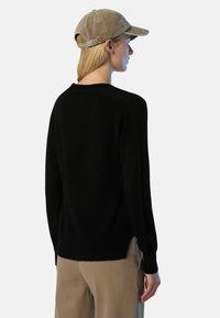 Frau mit einem schwarzen Langarmpullover, beigen Hosen und einer beigen Cord-Basecap, die leicht abgewandt steht.