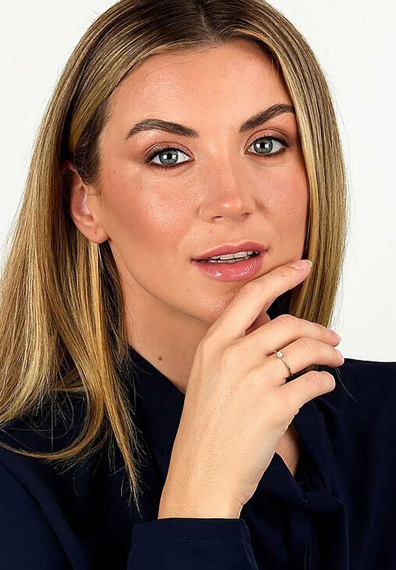 Nahaufnahme einer Person mit langen, glatten blonden Haaren, die ein dunkles Oberteil trägt und mit einem hellen Ring an ihrem Finger an ihrem Kinn spielt.