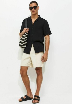 Uomo che indossa una camicia nera a maniche corte, pantaloncini beige, sandali neri e occhiali da sole, che tiene una borsa a righe bianco e nero.
