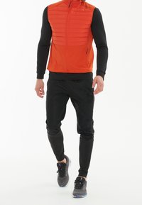Aktiv klädesset för män med en pufferväst i orange och svart långärmad tröja; kombinerat med svarta kroppsnära byxor och grå träningsskor.