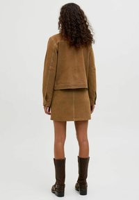 Veste en daim marron et jupe assortie, avec des détails de couture visibles. Cheveux bouclés, portant des bottes marron jusqu'aux genoux. Fond uni.