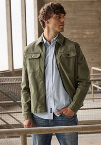 Olijfgroene jas met twee borstzakken, gedragen over een lichtblauw geruit overhemd, gecombineerd met een blauwe spijkerbroek. Gestileerd in een moderne, getailleerde pasvorm.