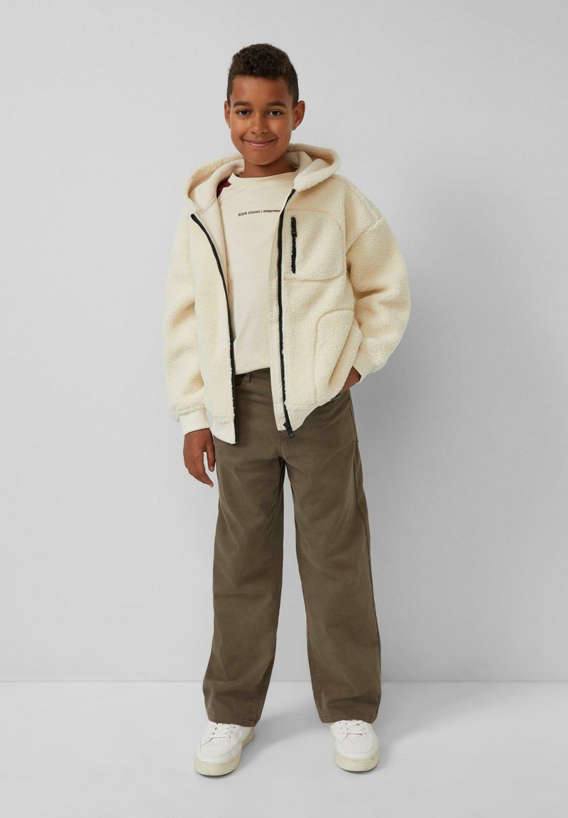 Fleecejas in crème, met een capuchon, een rits aan de voorkant en zijzakken. Gecombineerd met een lichtbeige shirt en een bruine broek. Sneakers in het wit.
