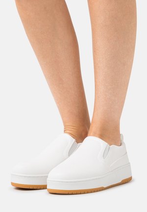 Sapatos brancos de slip-on com um cabedal de couro liso, sola branca grossa e base de borracha de cor goma. Apresentam detalhes elásticos nas laterais para facilitar o uso.