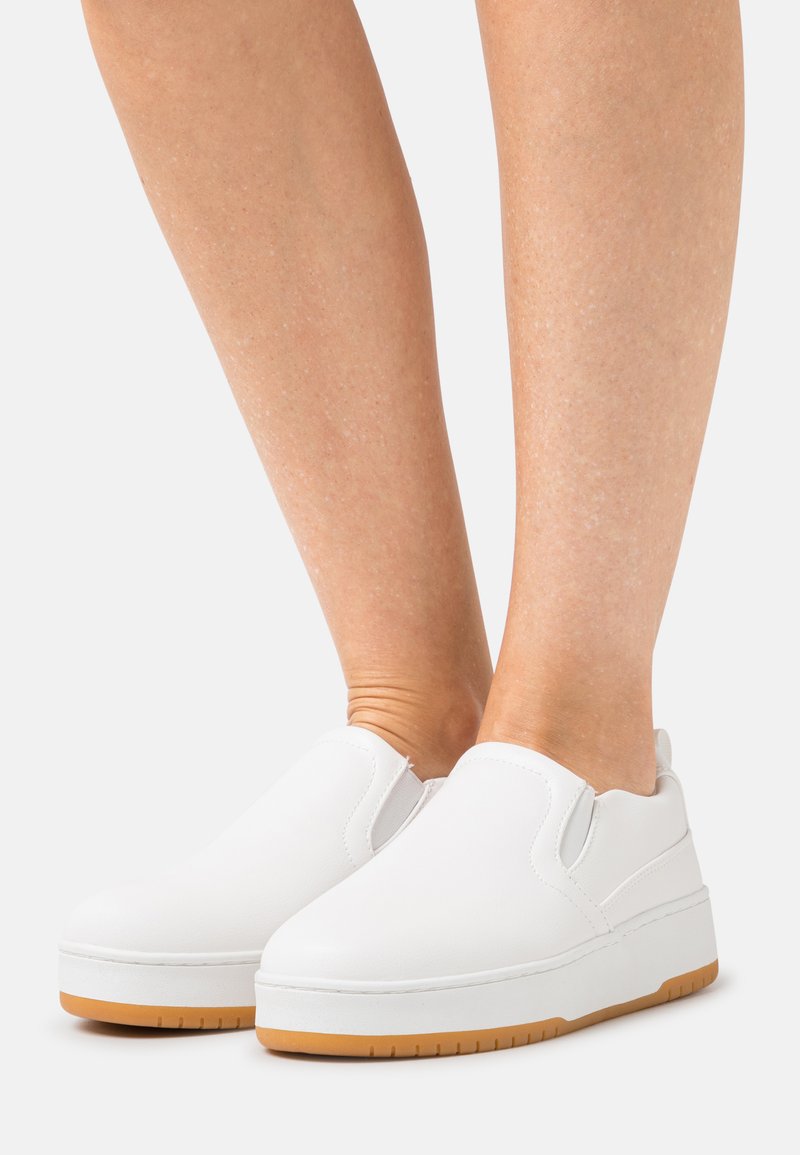 Sapatos brancos de slip-on com um cabedal de couro liso, sola branca grossa e base de borracha de cor goma. Apresentam detalhes elásticos nas laterais para facilitar o uso.