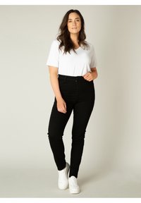 Zwarte high-waisted jeans met een slim fit, gecombineerd met een witte V-hals t-shirt. Model draagt witte sneakers, met golvend bruin haar.