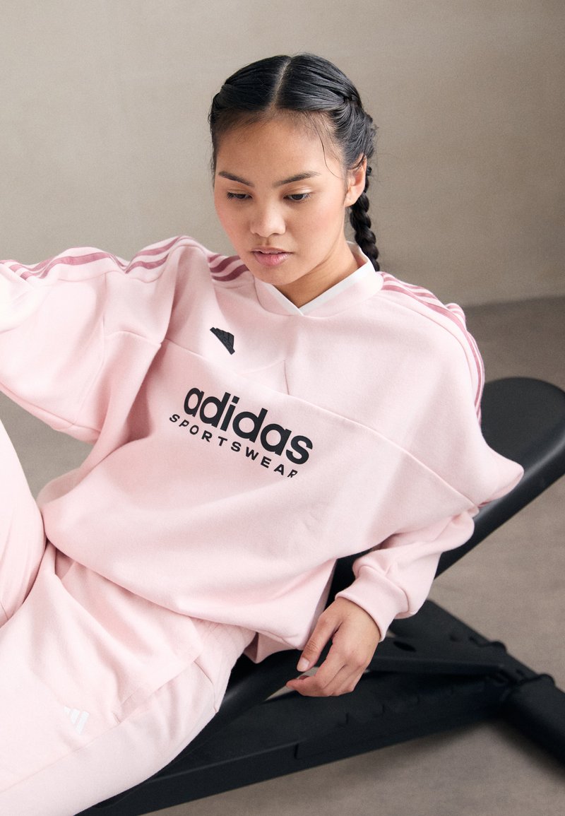 Jonge vrouw met gevlochten haar in roze Adidas sportkleding, liggend op een zwarte trainingbank in een neutrale binnenshuis omgeving.