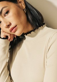 Haut beige côtelé avec un col montant, orné d'une bordure froncée noire. Le mannequin a les cheveux foncés jusqu'aux épaules et porte des boucles d'oreilles en or en forme de cercle.