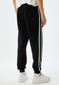 Pantalones de chándal negros de material suave con rayas laterales blancas. Cuentan con una cinturilla elástica y tobillos ajustados. Combinados con zapatillas blancas.