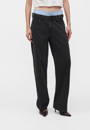 Personne portant un jean noir ample avec une taille visible à carreaux bleus et blancs et des chaussures à talons hauts pointues foncées, debout devant un fond blanc.