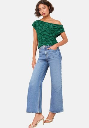 Grünes Blumen-Oberteil mit schulterfreiem Design aus strukturiertem Stoff, kombiniert mit hellblauen weit geschnittenen Jeans. Sandalen mit dünnem Riemen runden den Look ab.