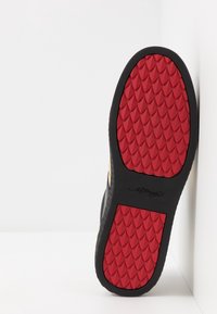Suela de zapato negra con una superficie de goma roja texturizada, que presenta un patrón de diamantes y un contorno negro. Detalles en dorado visibles en el lateral.