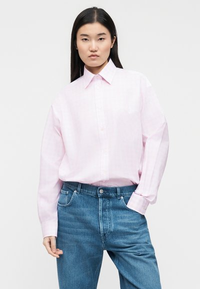 Γυναίκα με μακριά μαύρα μαλλιά, φορώντας χαλαρό ανοιχτό ροζ πουκάμισο με κουμπιά, περασμένο μέσα σε μπλε τζιν, μπροστά σε απλό λευκό φόντο.