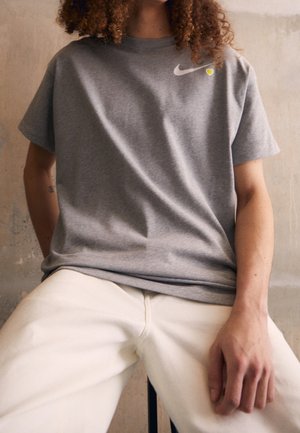 Personne aux cheveux bouclés portant un t-shirt Nike gris avec un petit cœur jaune et un pantalon crème, assise contre un mur uni.