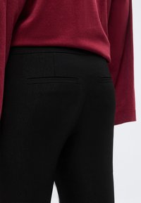 Κοντινό πλάνο ενός ατόμου που φοράει στενό μαύρο παντελόνι με πίσω τσέπες με ραφή και ένα σκουροκόκκινο μακρυμάνικο μπλουζάκι που είναι μπουσουλιασμένο μέσα.