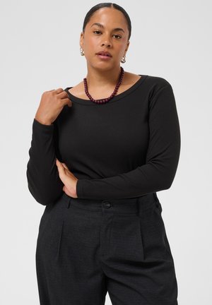 Femme aux cheveux foncés portant un haut noir à manches longues et un pantalon taille haute foncé, avec un collier de perles et des boucles d'oreilles anneaux, posant avec une main levée.