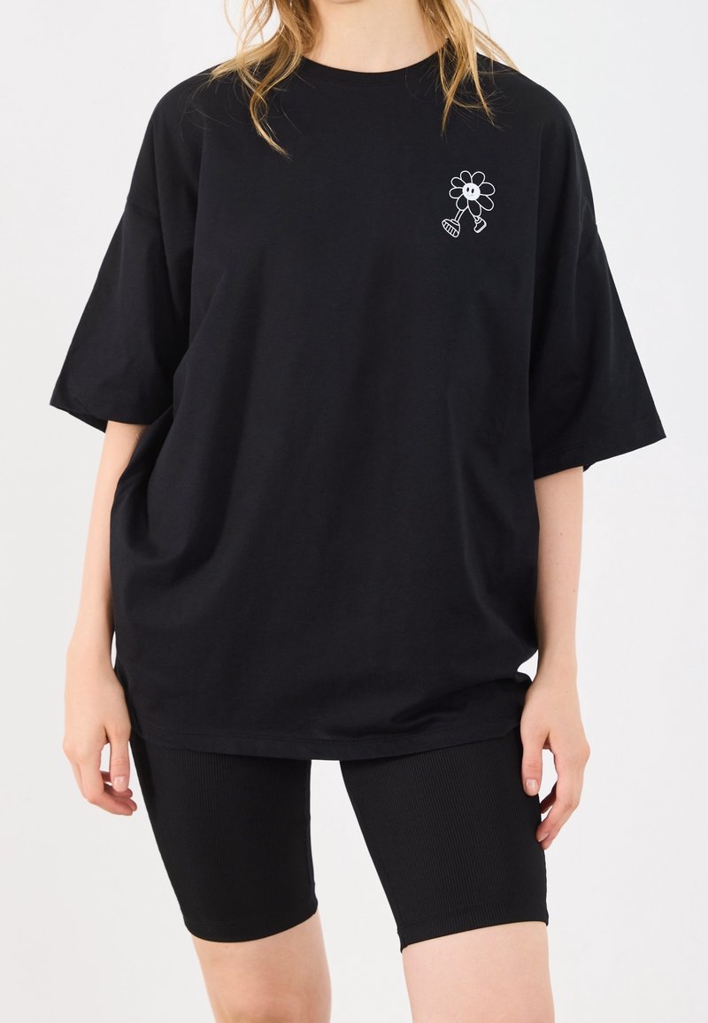 Personne portant un T-shirt noir surdimensionné avec un petit motif de fleur de cartoon blanche près de l'épaule gauche et un short noir côtelé.