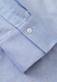 Polsino di camicia a quadri blu chiaro con un bottone bianco. Il tessuto presenta un motivo a griglia textured. Dettagli di cucitura pulita sui bordi.
