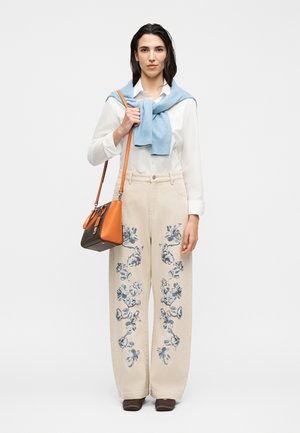 Femme portant une chemise blanche, un pantalon beige à imprimés floraux bleus, un pull bleu drapé sur les épaules, tenant un sac à main marron et orange.