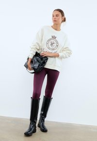 Felpa crema con logo bordeaux, abbinata a leggings viola scuri e stivali neri alti fino al ginocchio. La modella tiene una borsa nera.