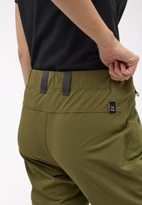 Osoba v olivově zelených kalhotách upravuje pásek vedle černého trička, na kalhotách je vidět logo Haglöfs.