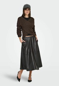 Pull en maille croisée brun foncé, associé à une longue jupe en cuir noir avec poches latérales. Des escarpins noirs pointus complètent le look.